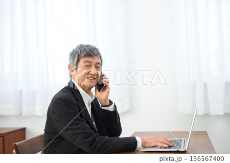 ノートパソコンを見ながらスマホで通話する60代男性 136567400