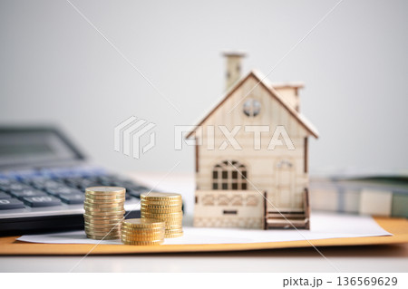 Miniature wooden house beside stacks of coins on paperwork with calculator in background, showing mortgage budgeting, real estate investment planning, long-term financial security. investment planning Miniature wooden house beside stacks of coins on paperwork with calculator in background, showing mortgage budgeting, real estate investment planning, long-term financial security. investment planning 136569629