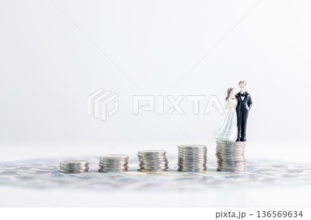 Bride and groom figurines standing on rising stacks of coins over banknotes with wide white copy space, suggesting wedding savings, budgeting and smart money management for financial growth. savings 136569634