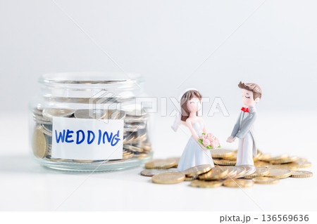 Jar labeled WEDDING filled with coins sits beside bride and groom figurines on scattered money over clean white background, representing saving, budgeting and financial planning for marriage. saving 136569636