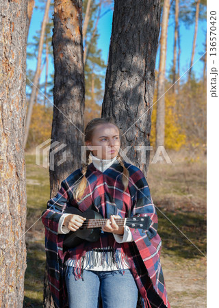 Beautiful blonde girl playing guitar ukulele and singing relaxing enjoying outdoors on sunny autumn day. Hippie woman in boho style training playing musical instrument. Fall leaves 136570420