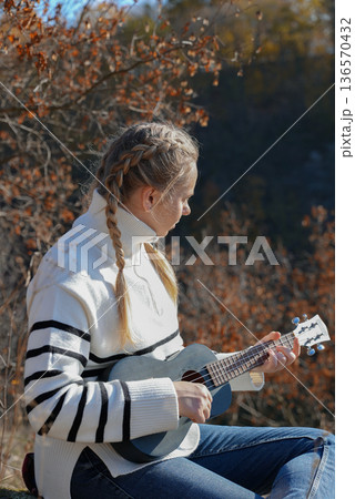 Beautiful blonde girl playing guitar ukulele and singing relaxing enjoying outdoors on sunny autumn day. Hippie woman in boho style training playing musical instrument. Fall leaves Beautiful blonde girl playing guitar ukulele and singing relaxing enjoying outdoors on sunny autumn day. Hippie woman in boho style training playing musical instrument. Fall leaves 136570432