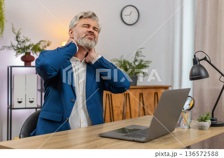 Middle-aged businessman grimaces sudden neck pain clutching neck seeking relief during work session 136572588
