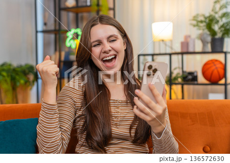 Young woman gets win alert on smartphone jumps and raises hand in victory and smiles big indoors yay 136572630
