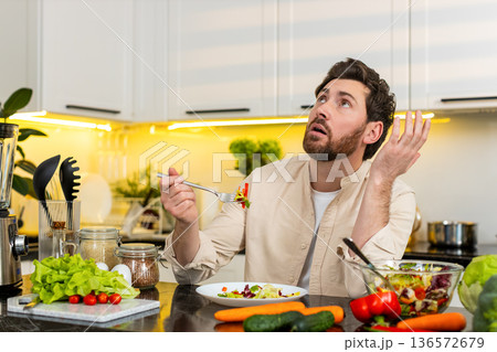 Middle-aged man unhappy eating fresh vegetable salad in home kitchen showing diet willpower struggle 136572679
