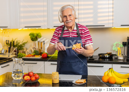 Senior old man dancing with headphones while making healthy breakfast toast for diet in home kitchen Senior old man dancing with headphones while making healthy breakfast toast for diet in home kitchen 136572816
