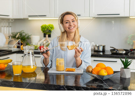 Young woman spreading peanut butter on toast while listening to music with headphones in own kitchen 136572896