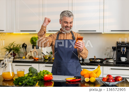 Middle-aged man celebrating big smartphone victory in home kitchen after online success notification 136572905