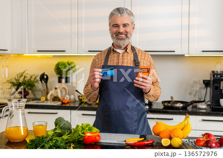 Middle-aged man uses smartphone with credit card in kitchen buying healthy groceries for diet plan Middle-aged man uses smartphone with credit card in kitchen buying healthy groceries for diet plan 136572976
