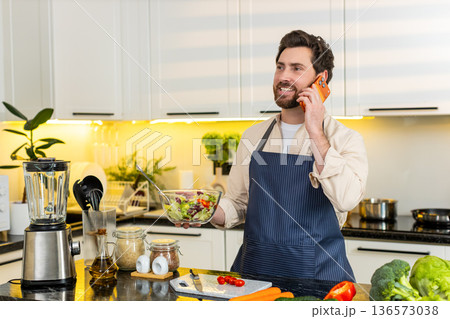Middle-aged man tasting fresh vegetable salad while talking on smartphone in home kitchen for diet Middle-aged man tasting fresh vegetable salad while talking on smartphone in home kitchen for diet 136573038