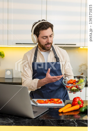 Middle-aged man in headphones making fit healthy salad for live stream cooking vlog in home kitchen 136573855
