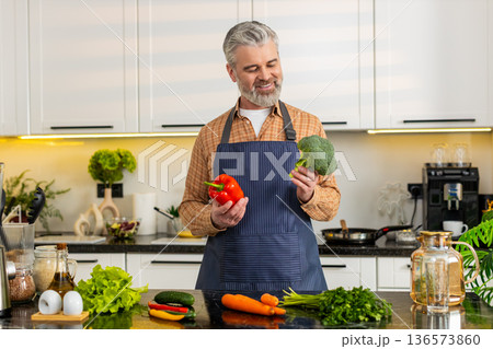 Middle-aged man smiling holding fresh pepper with broccoli for healthy diet prep at home kitchen fit 136573860