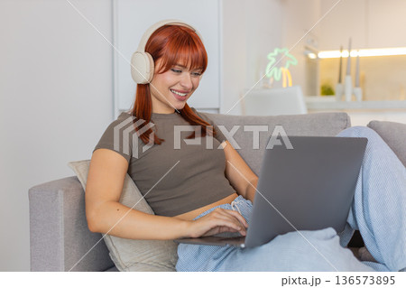 Young woman in headphones types email on laptop while music plays smiles and nods to rhythm focused Young woman in headphones types email on laptop while music plays smiles and nods to rhythm focused 136573895