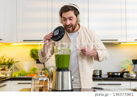 Middle-aged man excited wearing headphones showing green smoothie blender in home kitchen for diet 136573961