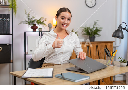 Young businesswoman closes laptop at end of day smiles shows thumbs up and exhales relief job done Young businesswoman closes laptop at end of day smiles shows thumbs up and exhales relief job done 136573966