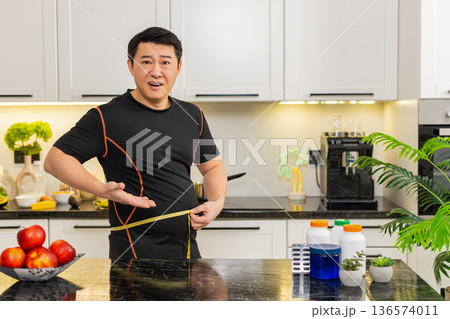 Heavy Asian adult man measures waist with tape upset about big belly fat weight gain at home kitchen Heavy Asian adult man measures waist with tape upset about big belly fat weight gain at home kitchen 136574011
