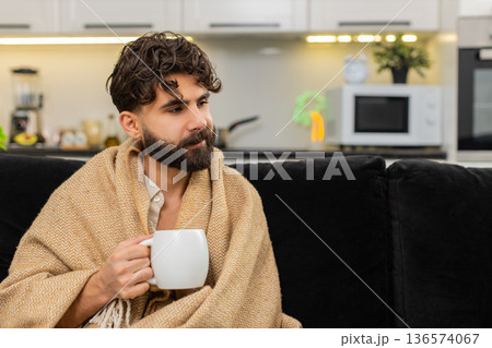 Indian man holding warm tea cup wrapped in blanket slowly sipping drink relaxing with gentle smile 136574067