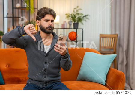 Indian man reads market drop news on smartphone loses game frowns thumbs down negative reaction mood Indian man reads market drop news on smartphone loses game frowns thumbs down negative reaction mood 136574079