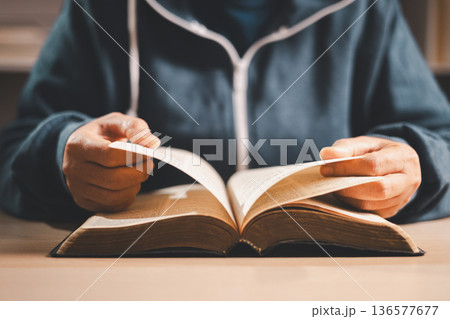 Man reading an open Bible at a desk, turning pages in warm indoor light with shallow depth of field, symbolizing Christian faith, devotion, study and quiet reflection at home. Man reading an open Bible at a desk, turning pages in warm indoor light with shallow depth of field, symbolizing Christian faith, devotion, study and quiet reflection at home. 136577677