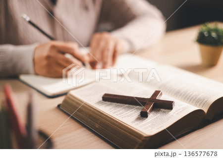 Person taking notes while reading an open Bible at a desk, pencil and journal beside a wooden cross, warm indoor light and soft focus, conveying Christian faith, devotion and scripture study at home. 136577678