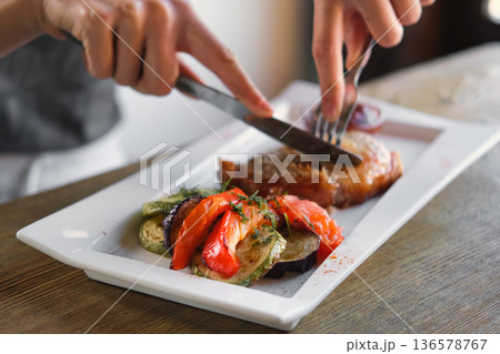 Girl cuts a cooked pork steak served on a white plate with grilled vegetables. Young woman using knife and fork to cutting meat eating in a restaurant. Balanced diet, cooking, culinary. Food concept Girl cuts a cooked pork steak served on a white plate with grilled vegetables. Young woman using knife and fork to cutting meat eating in a restaurant. Balanced diet, cooking, culinary. Food concept 136578767