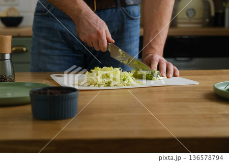 Handsome bearded man is preparing green fresh vegan salad in the kitchen at home. Male cutting vegetables, lettuce with knife on wooden chopping board, cooking vegetarian meal. Tasty healthy diet food Handsome bearded man is preparing green fresh vegan salad in the kitchen at home. Male cutting vegetables, lettuce with knife on wooden chopping board, cooking vegetarian meal. Tasty healthy diet food 136578794