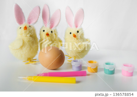 One Easter egg with bright colorful paints, markers and funny small toy chicks with Easter bunny ears on white background 136578854