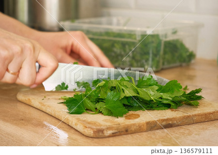 Person Chopping parsley bunch. Female cut greens with sharp knife at home kitchen. Woman hands cutting fresh aromatic herbs for cooking. Preparing food. Healthy food ingredient. Ecological products. 136579111