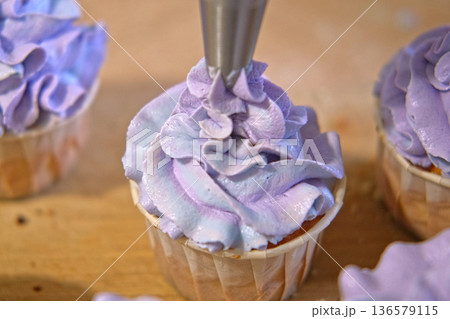 Person decorating cupcakes. Baker piping swirl of a creamy blue purple frosting onto a freshly baked cupcake. Decorated cakes with colorful sprinkles. Sweet Dessert Preparation. Pastry small business Person decorating cupcakes. Baker piping swirl of a creamy blue purple frosting onto a freshly baked cupcake. Decorated cakes with colorful sprinkles. Sweet Dessert Preparation. Pastry small business 136579115