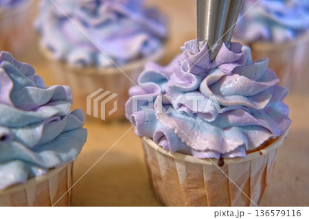 Person decorating cupcakes. Baker piping swirl of a creamy blue purple frosting onto a freshly baked cupcake. Decorated cakes with colorful sprinkles. Sweet Dessert Preparation. Pastry small business Person decorating cupcakes. Baker piping swirl of a creamy blue purple frosting onto a freshly baked cupcake. Decorated cakes with colorful sprinkles. Sweet Dessert Preparation. Pastry small business 136579116