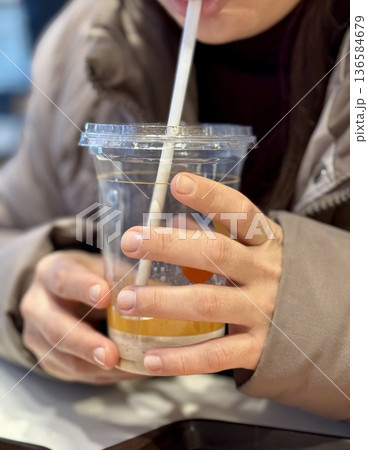 Hands Holding Iced Coffee Cup CloseUp, Student Gripping Cold Beverage With Both Hands At Cafe Table, Straw Near Lips, Condensation Beading On Plastic Cup, Casual Winter Jacket, Smartphone 136584679