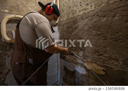 A man uses a hammer drill to remove old tiles from the walls. A construction worker, wearing ear protection and a respirator, uses a hammer drill to remove old plaster and cement from the wall. A man uses a hammer drill to remove old tiles from the walls. A construction worker, wearing ear protection and a respirator, uses a hammer drill to remove old plaster and cement from the wall. 136590886