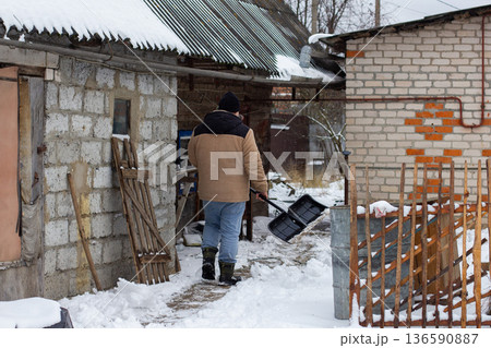 Man in winter clothing and hat walking on a snowy path, carrying a black snow shovel, preparing to clear accumulated snow from around residential brick buildings on a cold winter day. 136590887