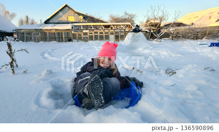 A happy young girl sits on a blue sled in deep, fresh snow, wearing a pink hat and warm clothes. The sweet child laughs and enjoys winter fun outdoors near her home. A happy young girl sits on a blue sled in deep, fresh snow, wearing a pink hat and warm clothes. The sweet child laughs and enjoys winter fun outdoors near her home. 136590896