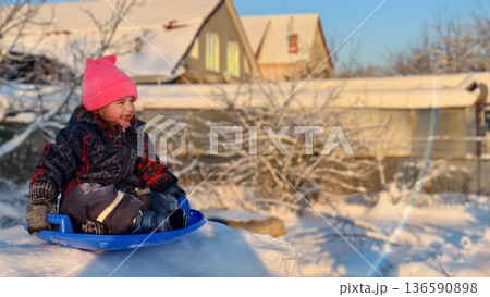 Young child experiencing joy and outdoor fun while actively sledging on fresh snow, wearing warm winter clothing, enjoying a sunny winter day with golden light. 136590898