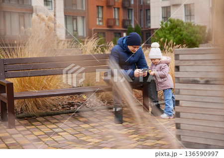 Father and daughter interacting with mobile phones while sitting on a bench in an urban park Father and daughter interacting with mobile phones while sitting on a bench in an urban park 136590997