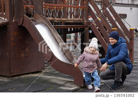 Father catching his toddler daughter sliding down a playground slide 136591002