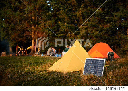 Solar panel in grassy field with tents in background highlights eco-friendly camping setup. Scene includes campers, emphasizing integration of renewable energy in outdoor activities. Solar panel in grassy field with tents in background highlights eco-friendly camping setup. Scene includes campers, emphasizing integration of renewable energy in outdoor activities. 136591165
