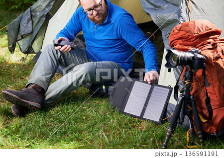 Bearded man sits at entrance of tourist tent in grassy area, charging power bank with portable solar panel. Nearby, tripod-mounted camera and orange backpack indicate his interest in photography. 136591191
