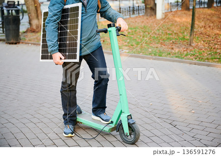 Cropped view of man rides electric scooter while carrying solar panel. Integration of solar power as sustainable energy source for charging electric scooters, promoting eco-friendly transportation. Cropped view of man rides electric scooter while carrying solar panel. Integration of solar power as sustainable energy source for charging electric scooters, promoting eco-friendly transportation. 136591276