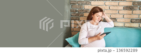 Pregnant caucasian female relaxing on sofa using tablet, brick wall background 136592289