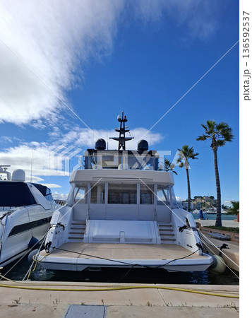 A large yacht is docked at the marina. The sun shines brightly in the sky. Palm trees stand tall around the area. Other boats are visible in the background. The scene shows a busy waterfront. 136592537