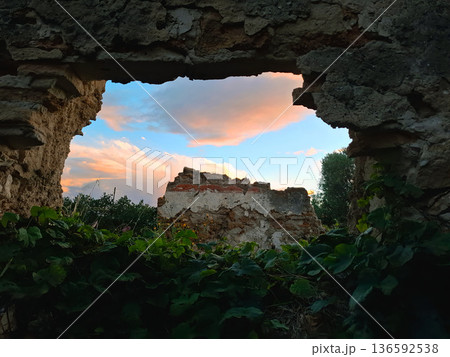 A broken wall shows a view of ruins and the sky with colorful clouds. The plants grow around the wall in the foreground. The scene is in the evening. A broken wall shows a view of ruins and the sky with colorful clouds. The plants grow around the wall in the foreground. The scene is in the evening. 136592538