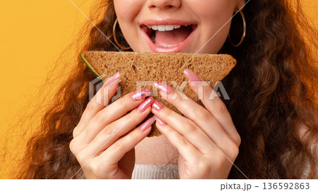 Woman smiles while holding a sandwich in front of her mouth against a yellow background 136592863