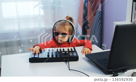 Cute little girl plays midi keyboard at home wearing headphones. She enjoys crafting music in a bright room with colorful decor 136592930