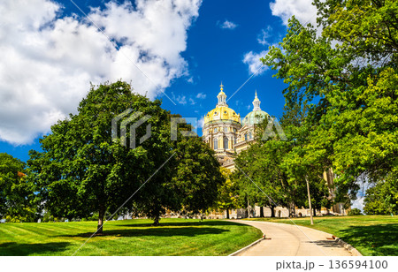 Iowa State Capitol in Des Moines. Historic architecture features the central golden dome and smaller copper domes viewed through green trees under a blue sky 136594100
