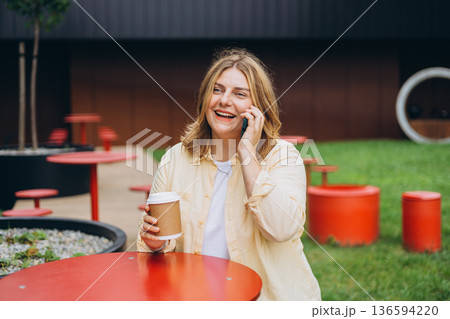 Happy 30s Woman Phone Call Cafe - Smiling woman talking on her phone while sitting at a table outside a cafe. 136594220