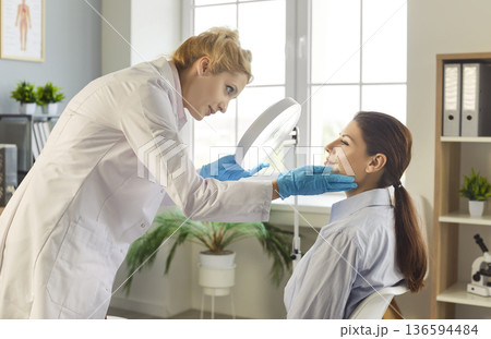 Beautician doctor, cosmetologist medical professional examining female face using magnifying mirror Beautician doctor, cosmetologist medical professional examining female face using magnifying mirror 136594484