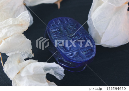 Lapis lazuli scarab base with engraved ancient egyptian hieroglyphs on dark stone with white petals. 136594630