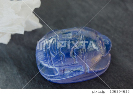 Lapis lazuli scarab figurine on dark stone with white petals, ancient Egyptian protection symbol. Lapis lazuli scarab figurine on dark stone with white petals, ancient Egyptian protection symbol. 136594633